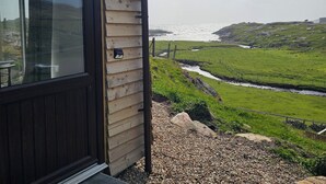 A wooden cabin with a large window, a gravel pathway leading to the door, and a view of a river and hills in the distance. - Oldshoremore Croft Cabins (Oldshore)