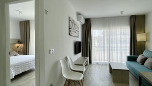 A modern hotel room with a bed, a desk with chairs, a television, a sofa, and a large window with curtains.
