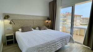 A hotel room with a large bed, a nightstand, a wall-mounted lamp, and a view of the balcony and buildings outside.