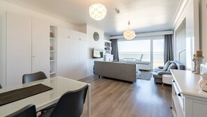 Ein modernes Wohnzimmer mit Essbereich, einer Couch und einem Fernseher. Ein großes Fenster bietet Meerblick.