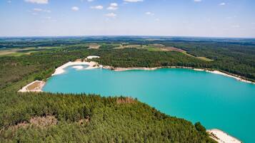 Une vue aérienne d’un grand lac turquoise, entouré d’une forêt dense, avec une petite plage et des champs situés au loin.