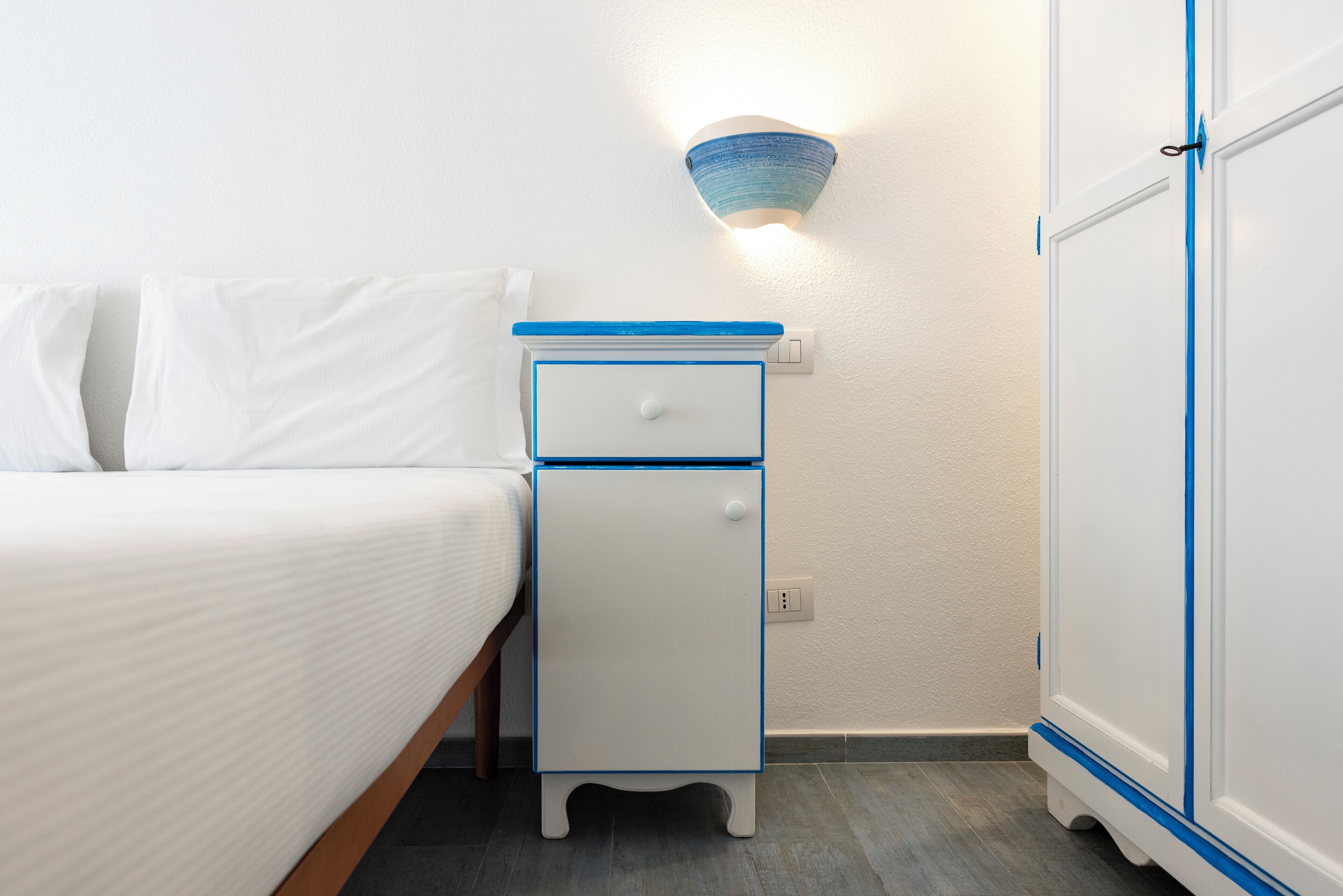 A neatly made bed with white linens, a white bedside table with blue trim, a wall-mounted light fixture, and a wardrobe with blue trim.
