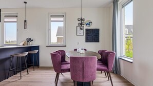 A modern kitchen with a dining area, featuring a round table and purple chairs, a bar area with stools, and large windows providing natural light. - Holiday Home in Medemblik near IJsselmeer (PS Opperdoes)