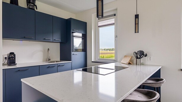 A modern kitchen with dark blue cabinets, a large island with a built-in stove, and a window offering a view of the outdoors. - Holiday Home in Medemblik near IJsselmeer (PS Opperdoes)
