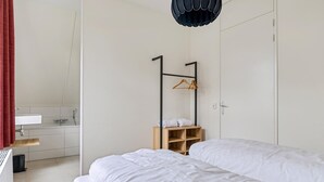 A modern hotel room with two beds, a wooden shelf with towels, a black hanging light fixture, and a view of the bathroom. - Holiday Home in Medemblik near IJsselmeer (PS Opperdoes)