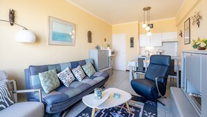 En moderne stue med sofa, lænestol og sofabord. Et køkken kan ses i baggrunden med hvide skabe og et spiseområde.