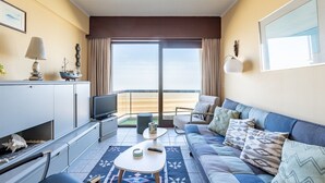 En moderne stue med sofa, sofabord og fjernsyn. Der kan ses en balkon med udsigt gennem glasdøren.
