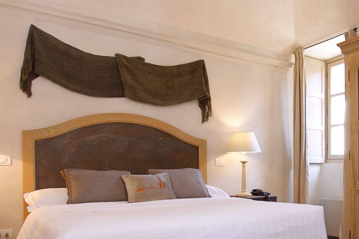 A large bed with a wooden frame and headboard, a bedside table with a lamp, and a window with curtains.