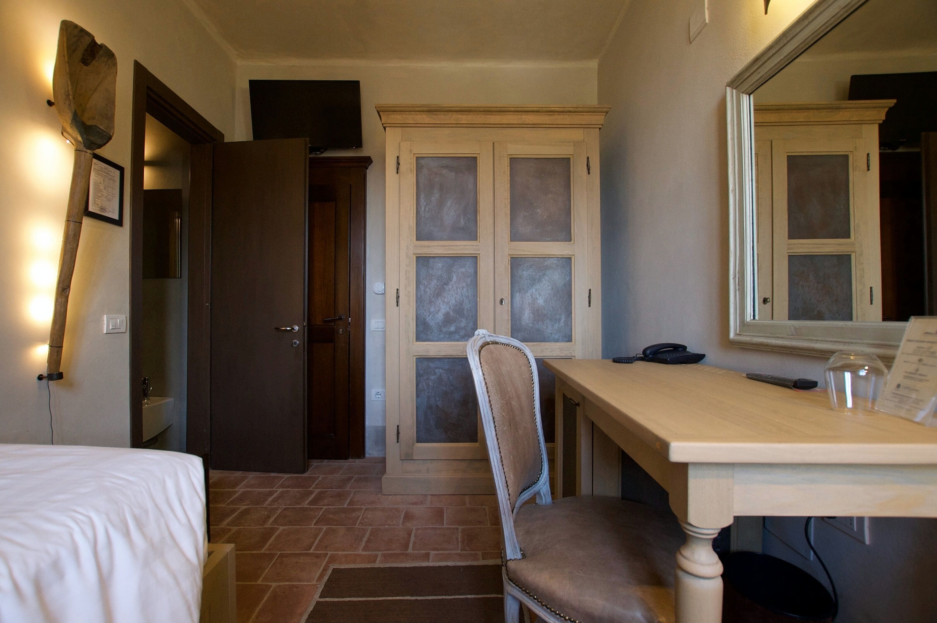 A hotel room with a bed, a desk with a chair, a mirror, a wardrobe, and a door with frosted glass panels.