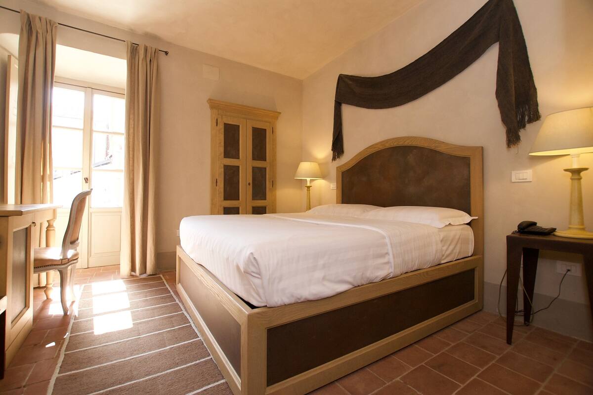 A hotel room with a bed, a desk, a chair, a lamp, and a window with curtains.