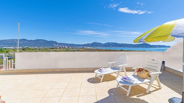 A balcony with two white chairs and a table, a yellow and white umbrella, and a view of the sea and mountains. - Apartment in Empuriabrava near Sandy Beach (Empuriabrava)