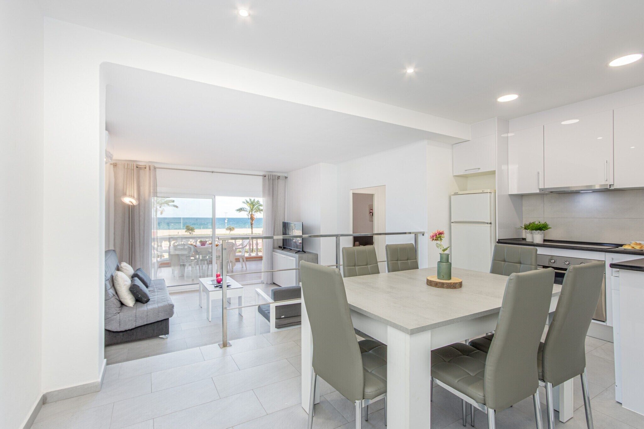 A modern kitchen with white cabinets, a dining area with a table and chairs, and a balcony with a view of the sea.