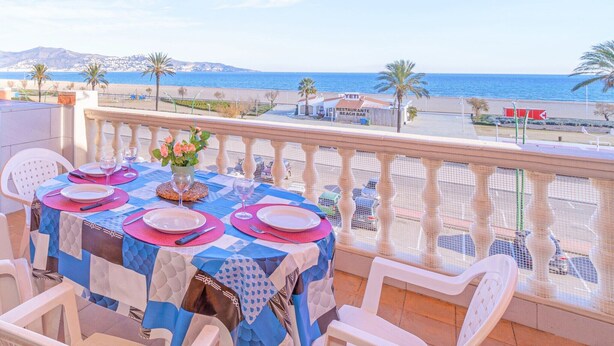 A balcony table set with plates, glasses, and a vase of flowers, overlooking a beach and a mountain in the background.