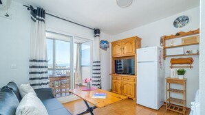 Un espace de vie compact comprenant un canapé, une table à manger, un coin cuisine avec des meubles en bois et un réfrigérateur, ainsi qu’un balcon avec des rideaux.