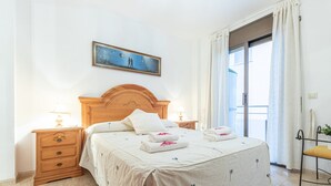 A neatly arranged bedroom with a wooden headboard, a bed with white linens, two bedside tables with lamps, a window with curtains, and a painting on the wall.