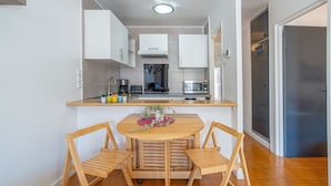 Une cuisine compacte avec des meubles blancs, un îlot central doté d’une table et de chaises, et un réfrigérateur placé à droite.