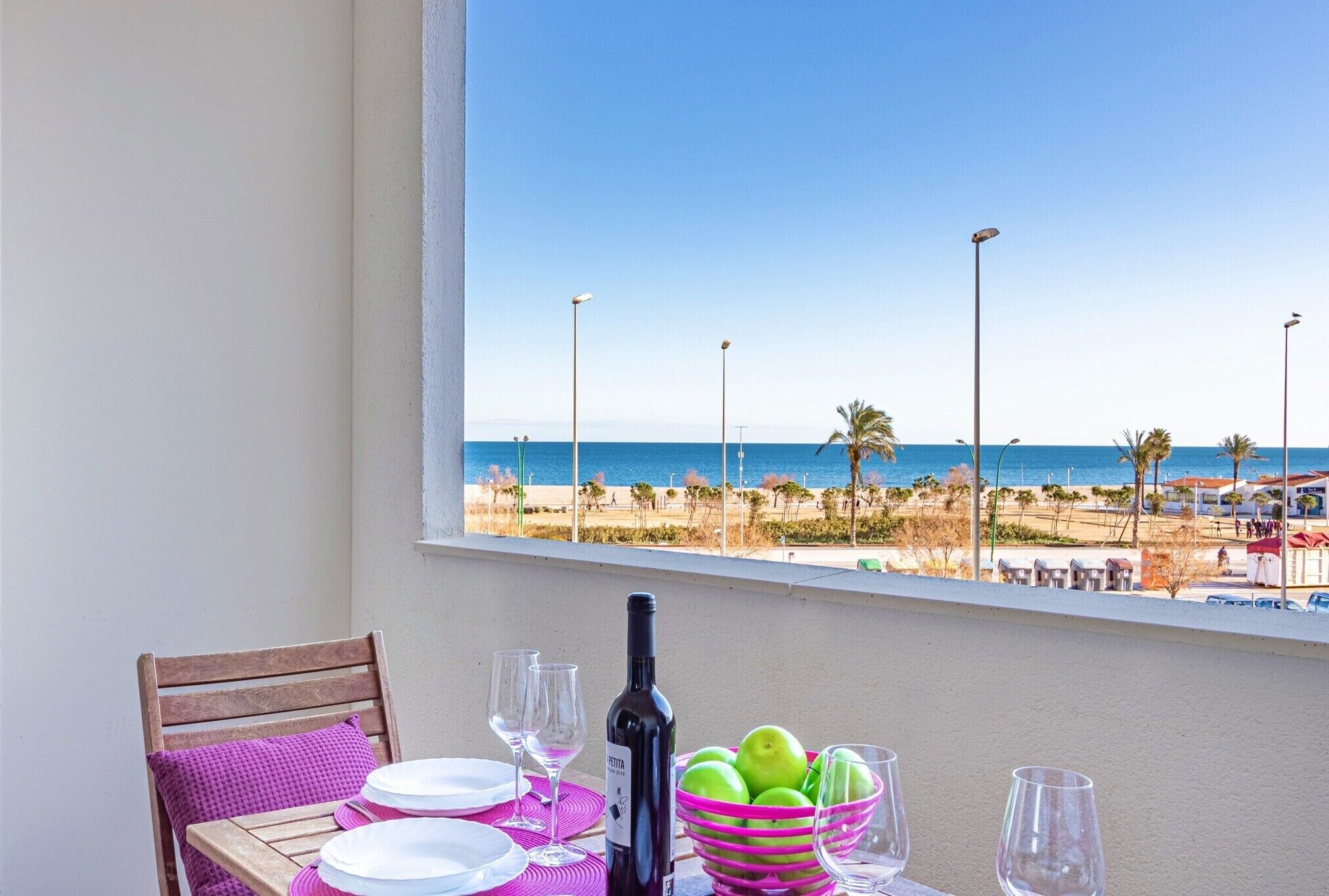 Une table dressée avec des assiettes, des verres à vin, une bouteille de vin et un panier de pommes vertes, sur un balcon offrant une vue sur la plage et l’océan.