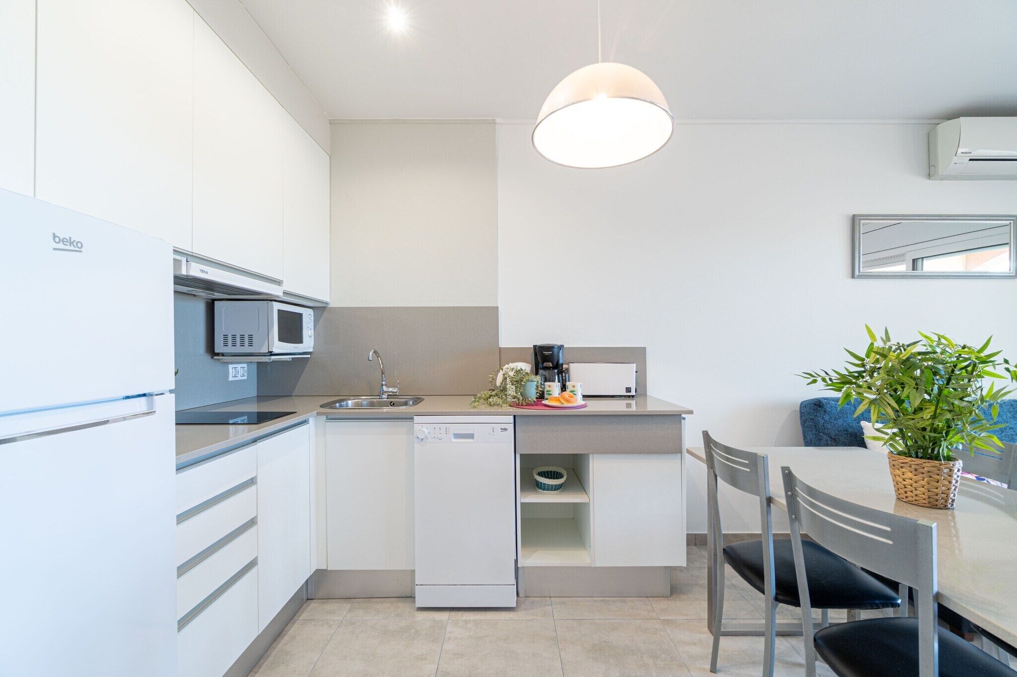 Une cuisine moderne avec des meubles blancs, un lave-vaisselle encastré, un four à micro-ondes et une machine à café. On y trouve une table à manger avec des chaises et une plante en pot posée sur la table.