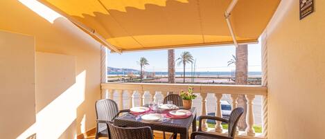 Un balcón con una mesa dispuesta para dos personas, con vistas a una playa con palmeras y un cielo despejado.