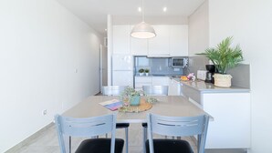 Een moderne keuken met witte kasten, een magnetron en een koffiezetapparaat. Er staat een eettafel met twee stoelen en een plant op het aanrecht.