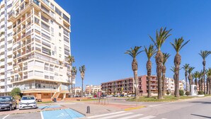 A multi-story residential building with balconies, a parking area, and palm trees.