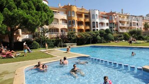 Una piscina rodeada de edificios de varios pisos con balcones, un área verde y gente relajándose.