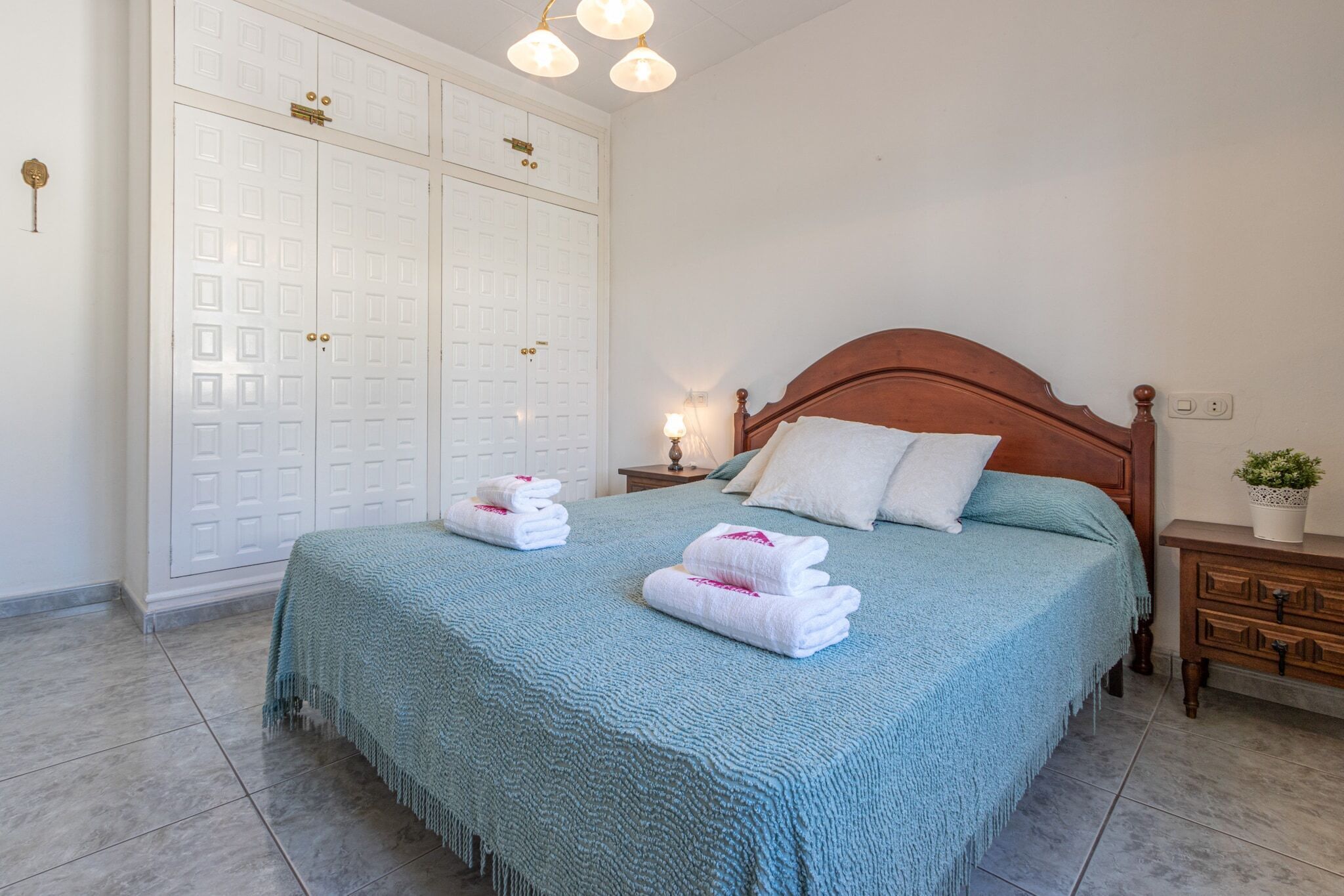 Een netjes opgemaakt bed met een blauwe spreidek en witte kussens, een houten nachtkastje met een lamp, en een witte kast met een sleutelgat.