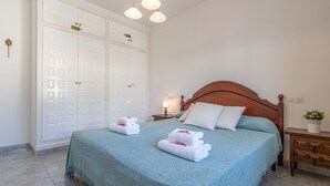 Ein ordentlich bezogenes Bett mit blauer Bettdecke und weißen Kissen, ein Nachttisch aus Holz mit einer Lampe sowie ein weißer Kleiderschrank mit Schlüsselloch.