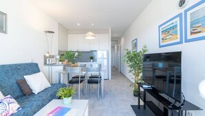 A modern living room with a sofa, coffee table, and TV. There is a dining area with chairs and a table, and a kitchen in the background. - Apartment in Empuriabrava by the Beach (Empuriabrava)