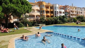 A swimming pool surrounded by multi-story buildings with balconies, a grassy area, and people relaxing.