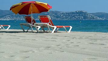 A beach with two red and white lounge chairs under a red and yellow umbrella, with a view of a coastal town and hills in the background.