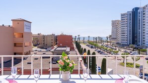 Un balcón con una mesa preparada para dos personas, con vista a un estacionamiento y edificios, y una vista despejada del mar.