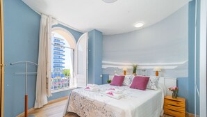 Un dormitorio con una cama grande, una ventana con cortinas, una mesita de noche con lámpara y vistas a un edificio.