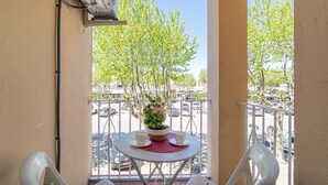 Un balcón con una mesa redonda, dos sillas, una planta en maceta y vistas a una calle con coches y árboles.
