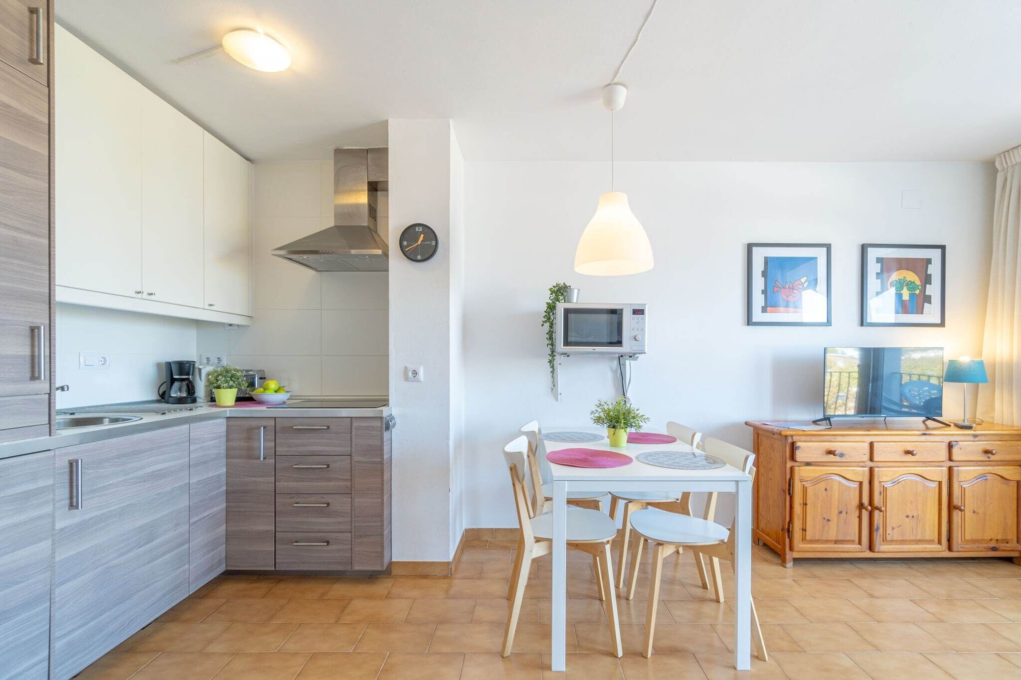 Een moderne keuken met witte kastjes, een eettafel met stoelen, een magnetron en een klok aan de muur.