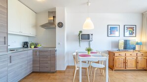 A modern kitchen with white cabinets, a dining table with chairs, a microwave, and a clock on the wall. - Apartment in Empuriabrava near Sandy Beach (Empuriabrava)
