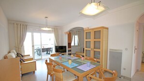 Un comedor con una mesa dispuesta para cuatro personas, rodeada de sillas de madera. Un televisor está montado en la pared, y hay un mueble grande con puertas de cristal a la derecha.