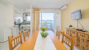 Un comedor con una mesa de madera larga, sillas y plantas en macetas. Se ve una cocina al fondo y un televisor montado en la pared.