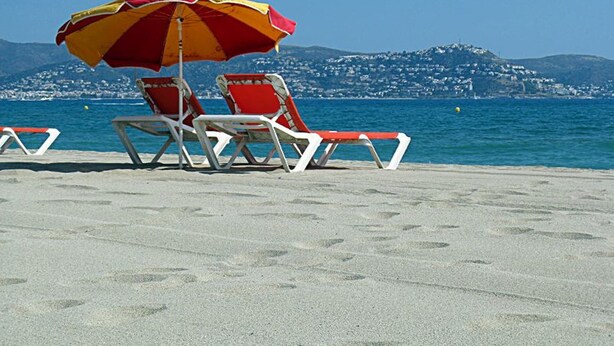 Una playa con dos tumbonas rojas y blancas bajo un paraguas rojo y amarillo, con una vista despejada del mar y una colina distante al fondo.