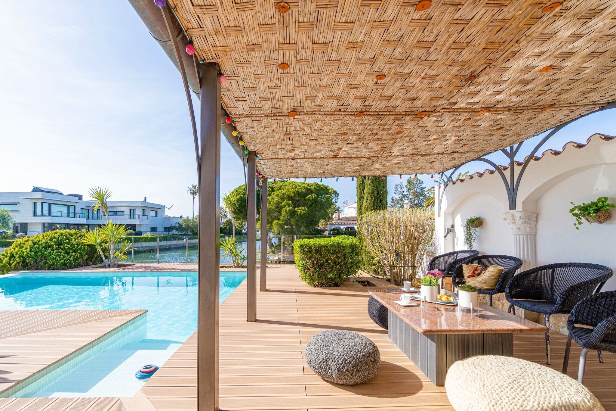 A poolside area with a wooden deck, a covered seating area with wicker chairs, and a view of the pool and surrounding greenery.