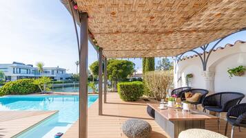 A poolside area with a wooden deck, a covered seating area with wicker chairs, and a view of the pool and surrounding greenery.