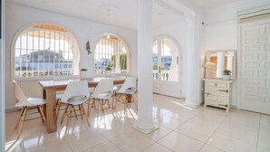 A spacious room with a wooden dining table, white chairs, and large arched windows offering a view of buildings outside. - Villa in Empuriabrava with Private Mooring (Empuriabrava)