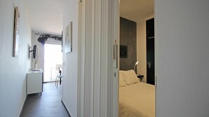 A narrow hallway leading to a bedroom with a bed and a bedside lamp. A desk with a chair is visible in the adjacent room.