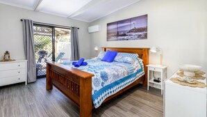 A bedroom with a wooden bed, a view of the sea through a large window, a white bedside table, and a wall-mounted air conditioner.