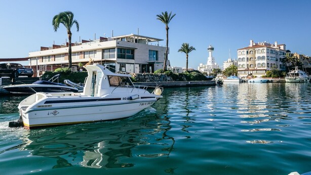 A marina with a white boat docked in the foreground, a modern building with balconies, and a lighthouse in the background. - Apartment in Empuriabrava with Canal Views (Empuriabrava)