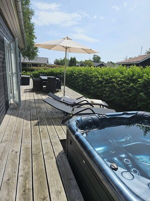 Een houten terras met een jacuzzi, lounge fauteuils en een parasol.