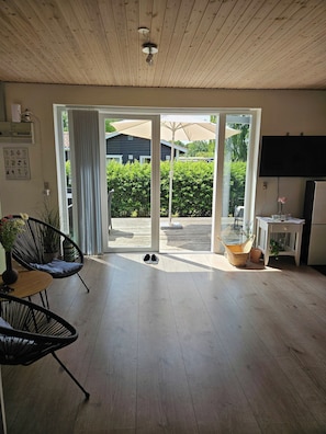 A spacious room with a wooden ceiling, a sliding glass door leading to an outdoor area, a flat-screen TV, and a small table with a vase.