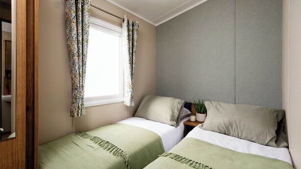 A bedroom with two beds, a window with floral curtains, a grey headboard, and a small potted plant on the nightstand. - Three bedroom caravan, ideal for families and angling groups. (Peterborough)