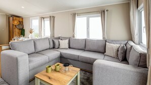 A modern living room with a grey sectional sofa, a wooden coffee table, and a view of the outdoors through large windows. - Three bedroom caravan, ideal for families and angling groups. (Peterborough)