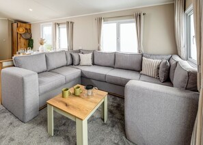 En moderne stue med en grå hjørnesofa, et træsofabord og udsigt til naturen gennem store vinduer.